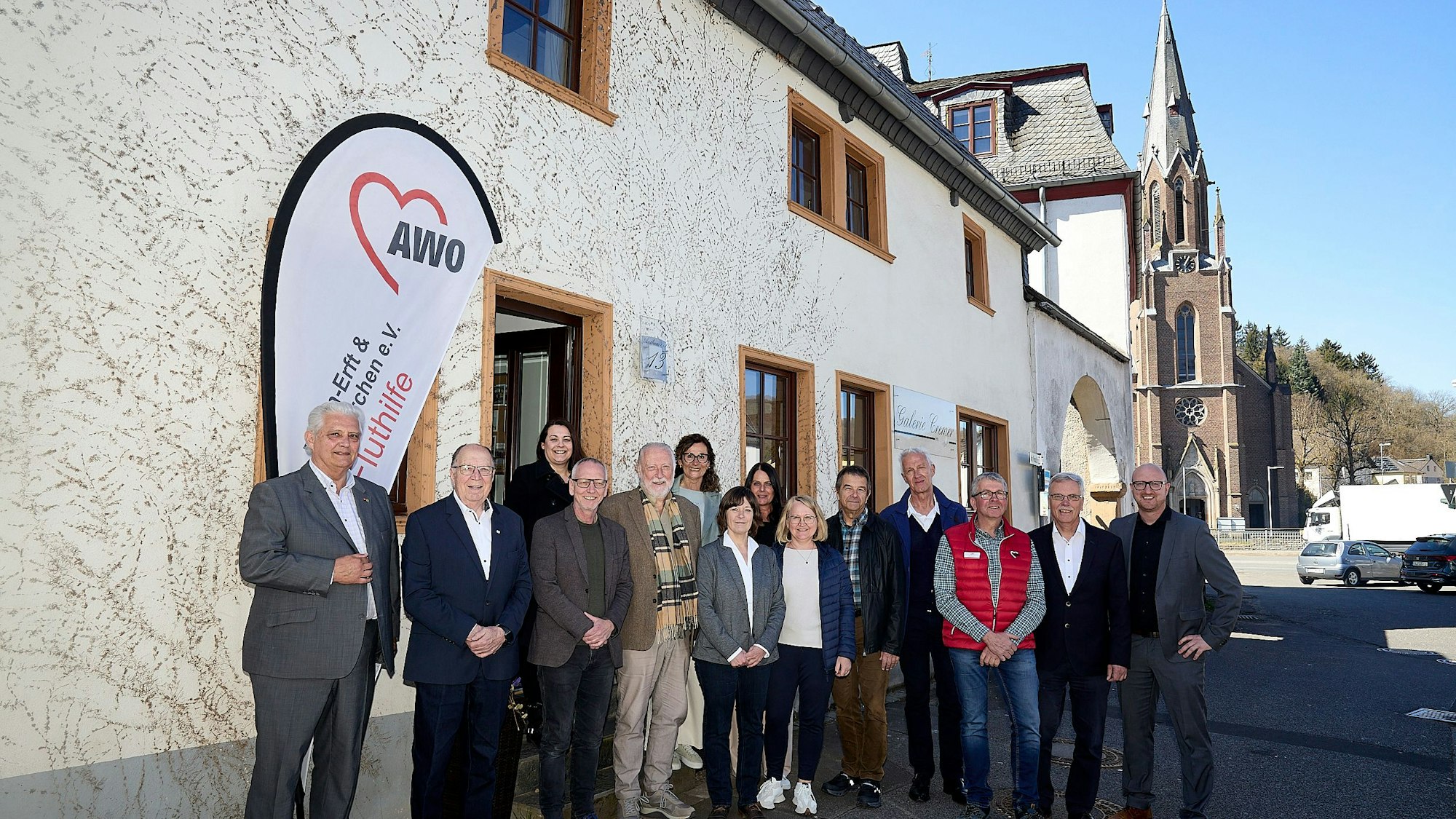 Mehrere Menschen stehen vor einem Haus. Im Hintergrund sind Fahrzeuge und eine Kirche.