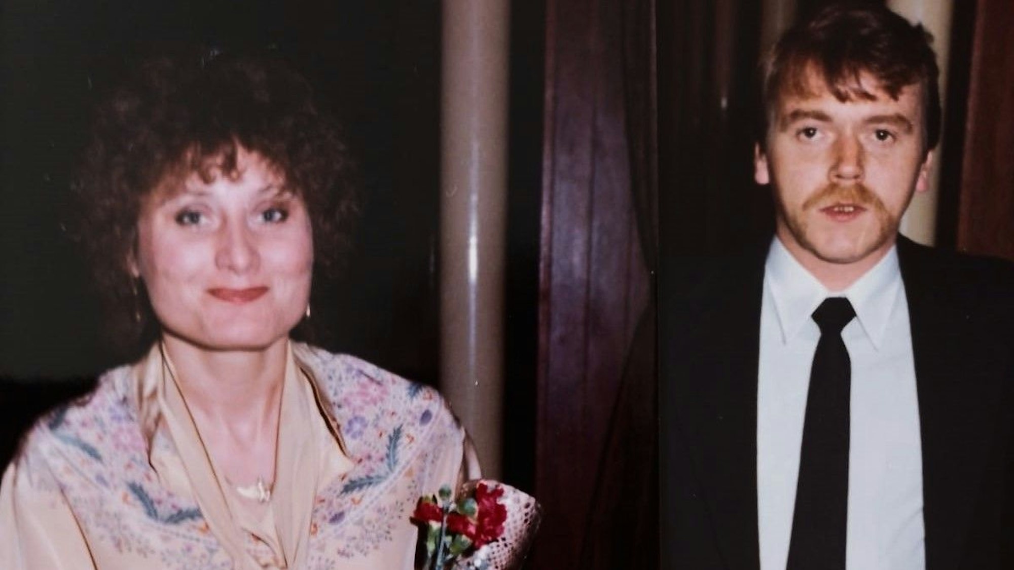 Ein Mann und eine Frau stehen vor einer dunklen Wand, die Frau hält Blumen.