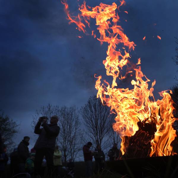 Ein Feuer brennt, drumherum sind Leute zu sehen, es ist Abend und im Hintergrund stehen Bäume.