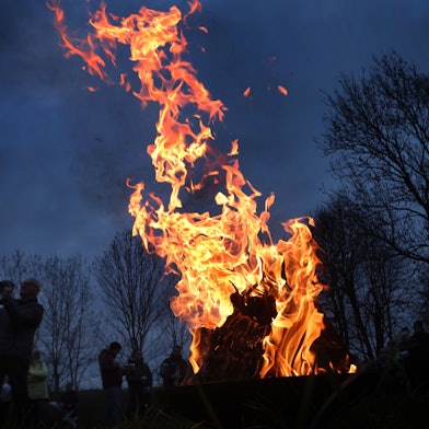 Ein Feuer brennt, drumherum sind Leute zu sehen, es ist Abend und im Hintergrund stehen Bäume.