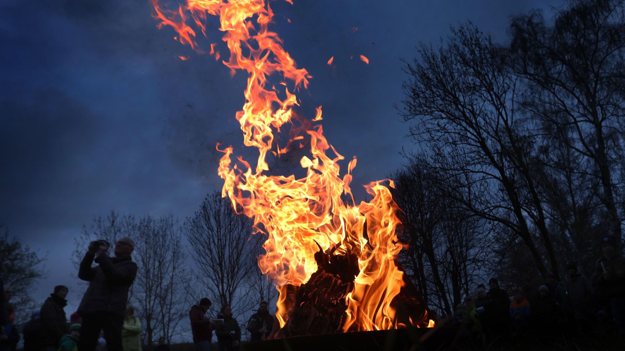 Ein Feuer brennt, drumherum sind Leute zu sehen, es ist Abend und im Hintergrund stehen Bäume.