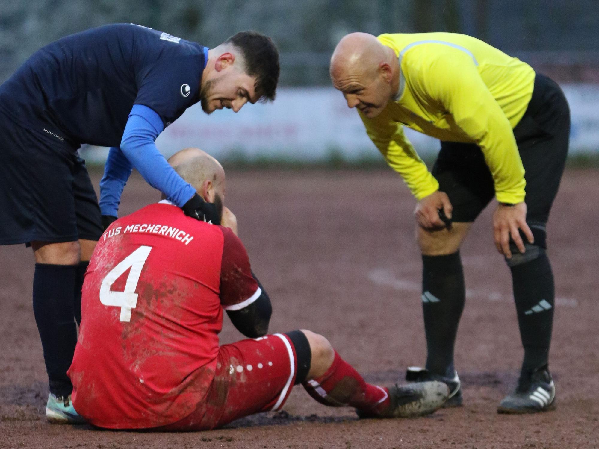 Das Bild zeigt Britz, der auf dem Boden sitzt. Bessenichs Spieler und der Schiedsrichter stehen bei ihm.