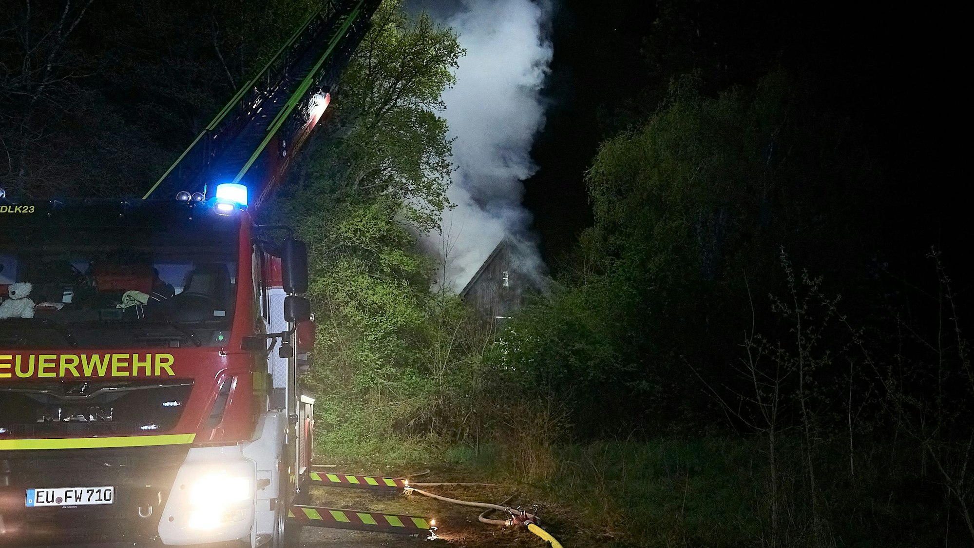 Die Drehleiter ist in Stellung gegangen. Im Hintergrund steigt eine Rauchwolke aus dem brennenden Gebäude.