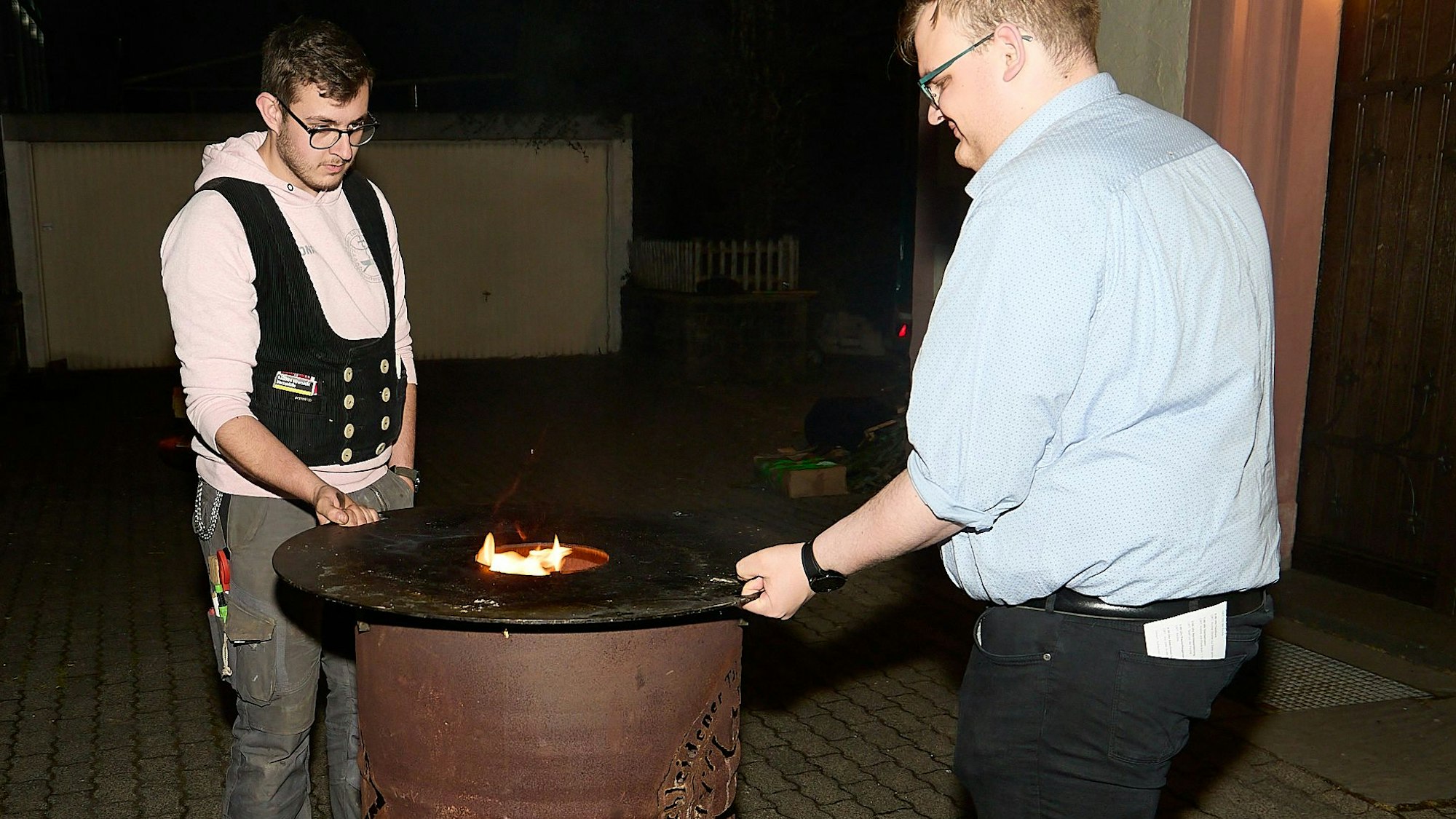 Zwei Teilnehmer legen einen gusseisernen Grill-Ring auf die brennende Feuertonne.