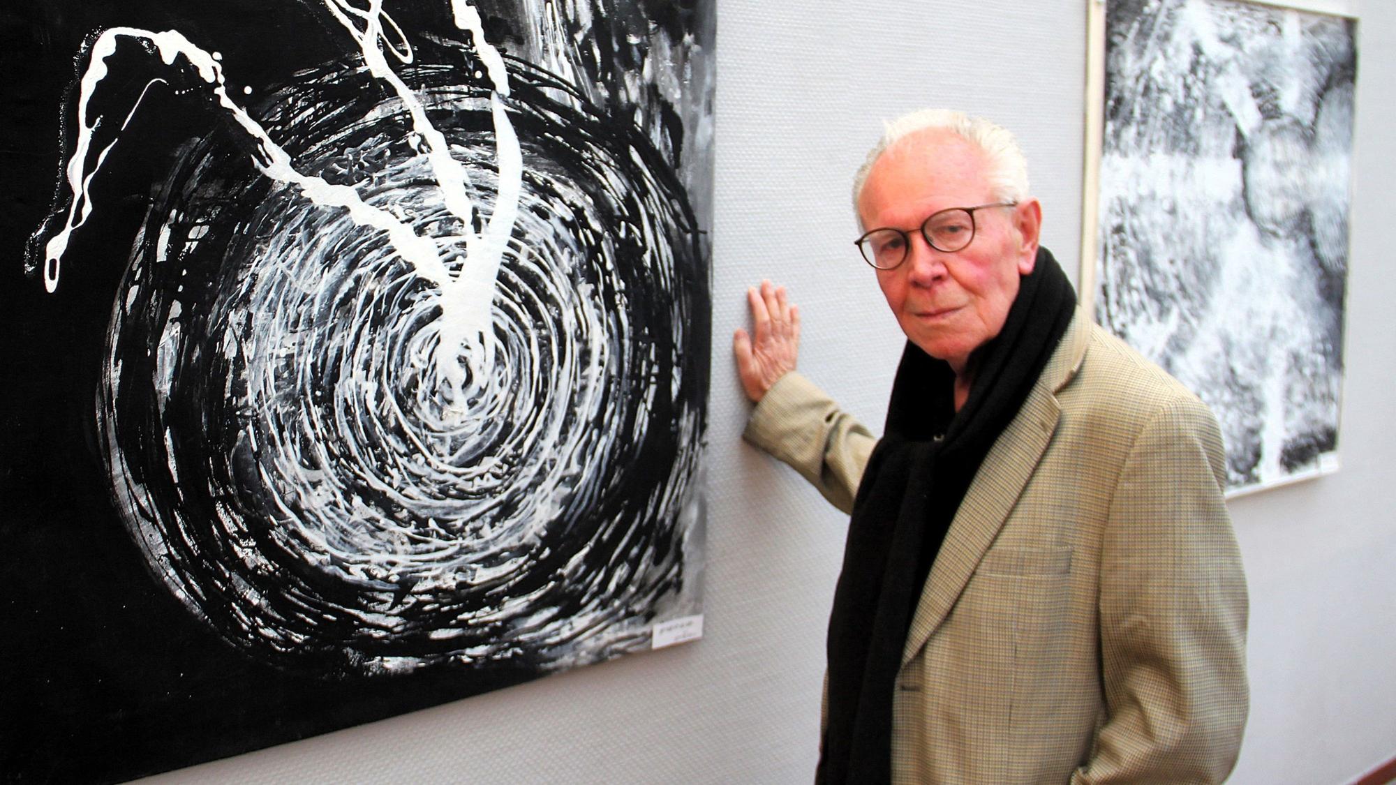 Ein älterer Herr steht in einer Galerie neben seinem abstrakten Schwarz-Weiß-Werk.
