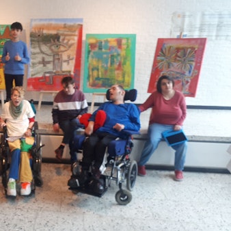 Menschen, teils im Rollstuhl sitzen vor einer Bühne, auf der fünf Bilder auf Staffleien stehen. Der Künstler, Mikail Akar, steht vor seinen Bildern.