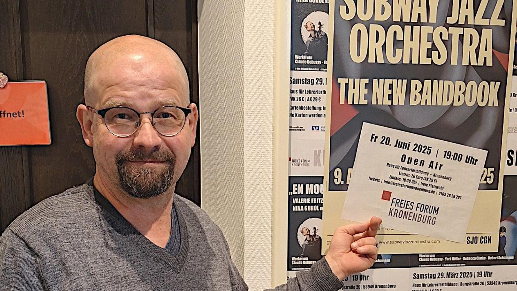 Martin Schöddert zeigt auf ein Plakat, auf dem „Subway Jazz Orchestra“ steht.