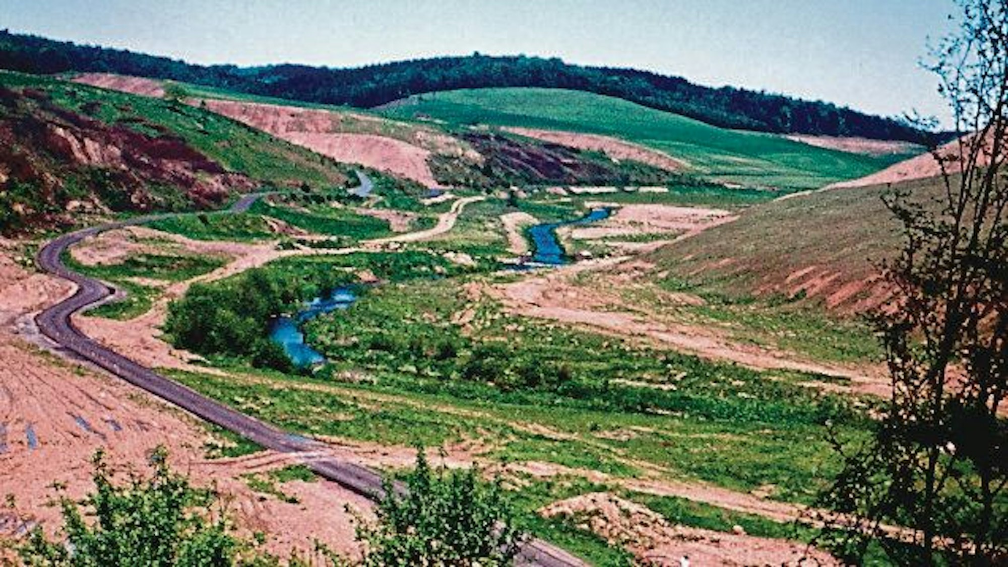 Durch ein leergeräumtes Tal ohne Bäume und Häuser fließt ein Bach.