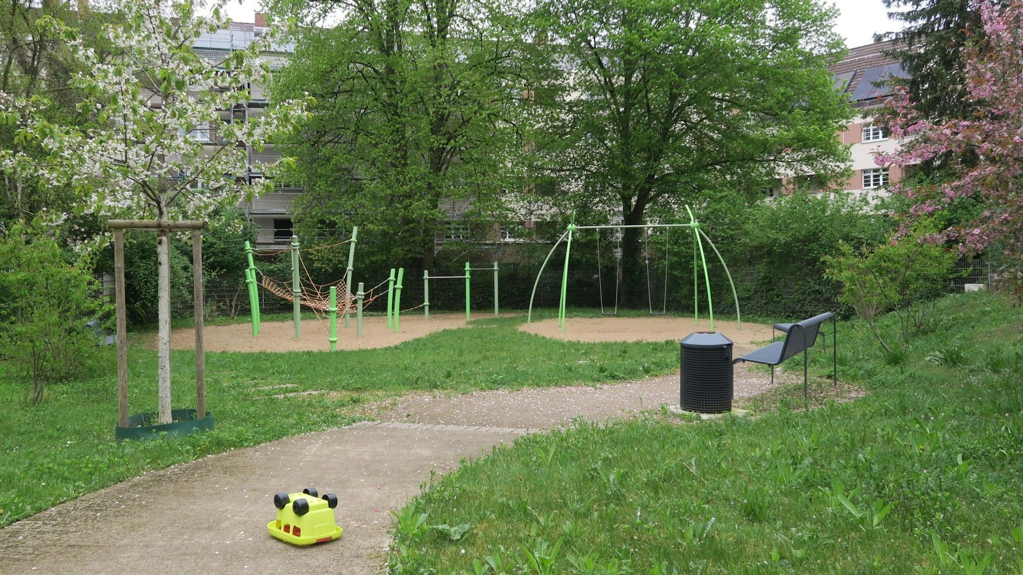 Ein grünes Spielgerüst steht auf einer Sandfläche, dahin führt ein asphaltierter Weg.