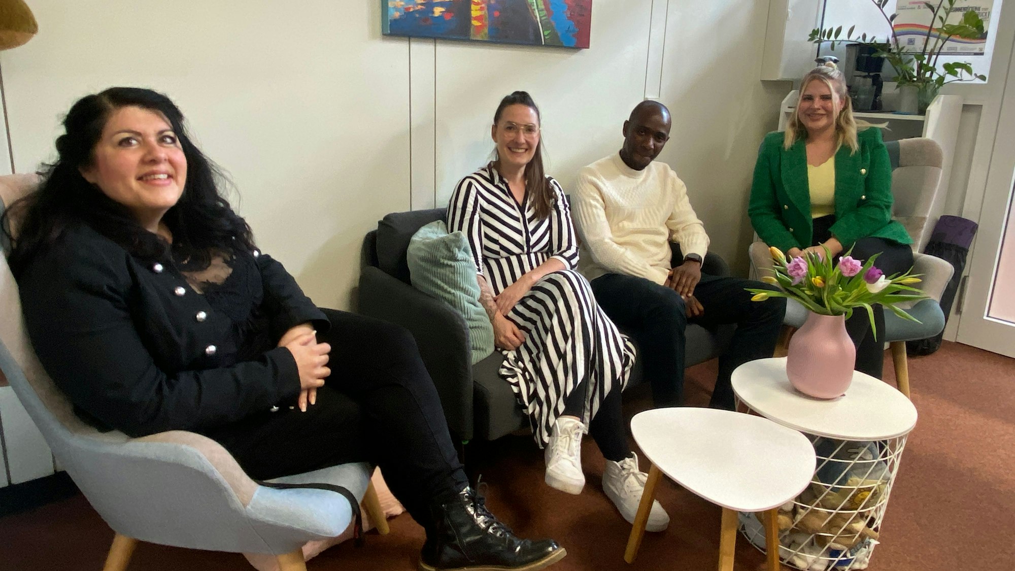 In einem Büro stehen ein Sofa und zwei Sessel, darauf sitzen drei Frauen und ein Mann. Auf einem Tischchen davor stehen Blumen.