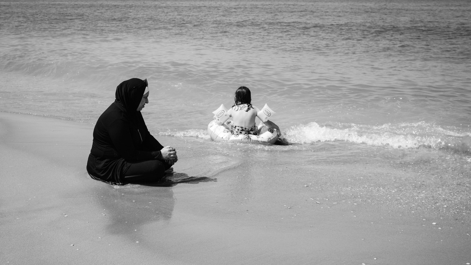 Ein schwarz-weiß-Bild zeigt eine verschleierte Frau, die am Meer sitzt. Daneben sitzt ein kleines Mädchen mit Schwimmring und Schwimmflügen im Wasser.
