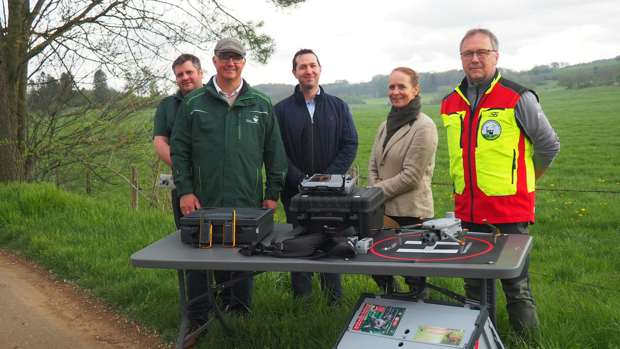 Vier Männer und eine Frau stehen auf einer Wiese hinter einem Tisch, auf dem eine Drohne und weitere technische Geräte liegen.