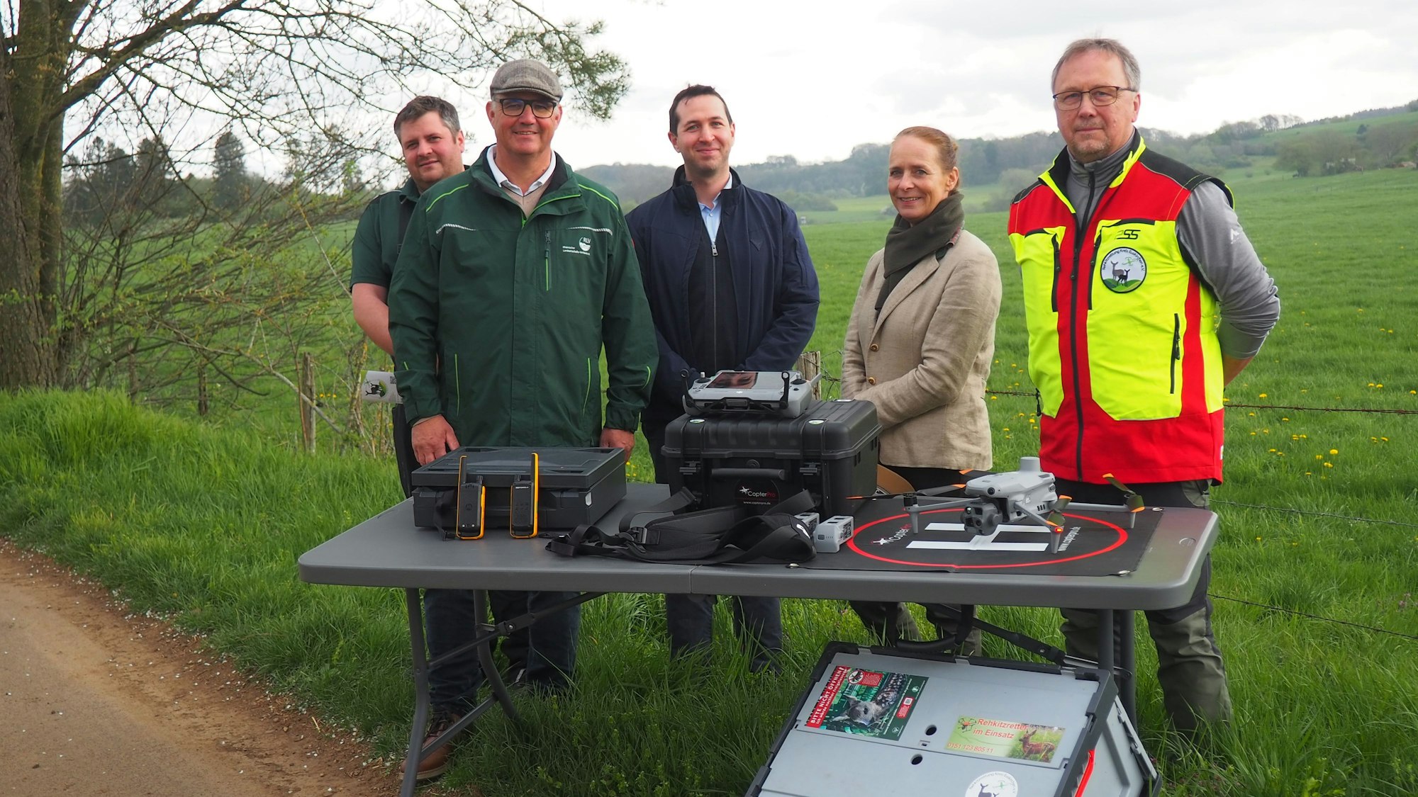 Vier Männer und eine Frau stehen am Wegesrand an einer Wiese. Vor ihnen ist ein Tisch aufgebaut, auf dem Koffer und eine Drohne liegen.