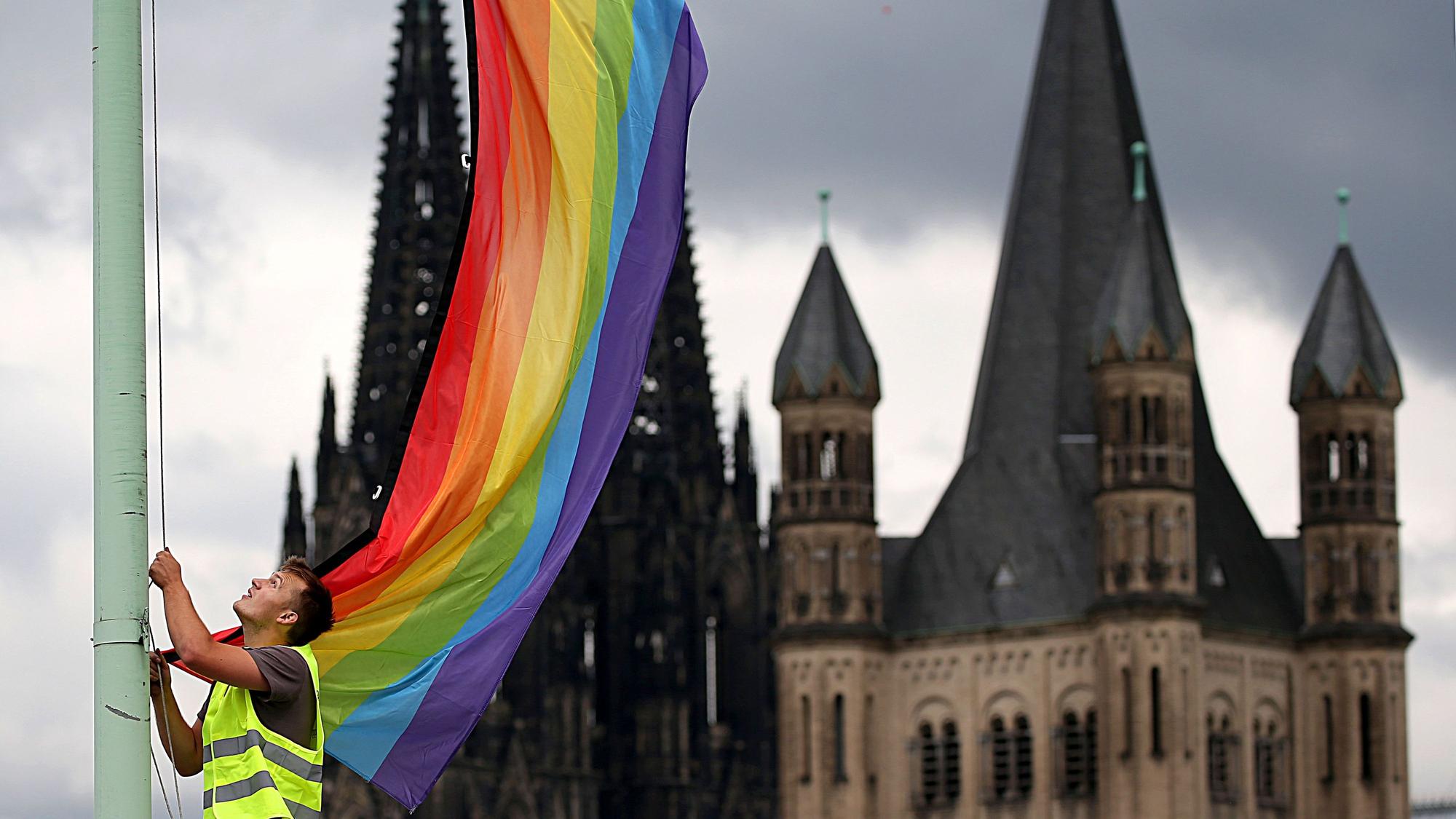 Köln: Ein Arbeiter hängt vor der Kulisse des Doms und der Kirche Groß St. Martin eine Regenbogenfahne auf.
