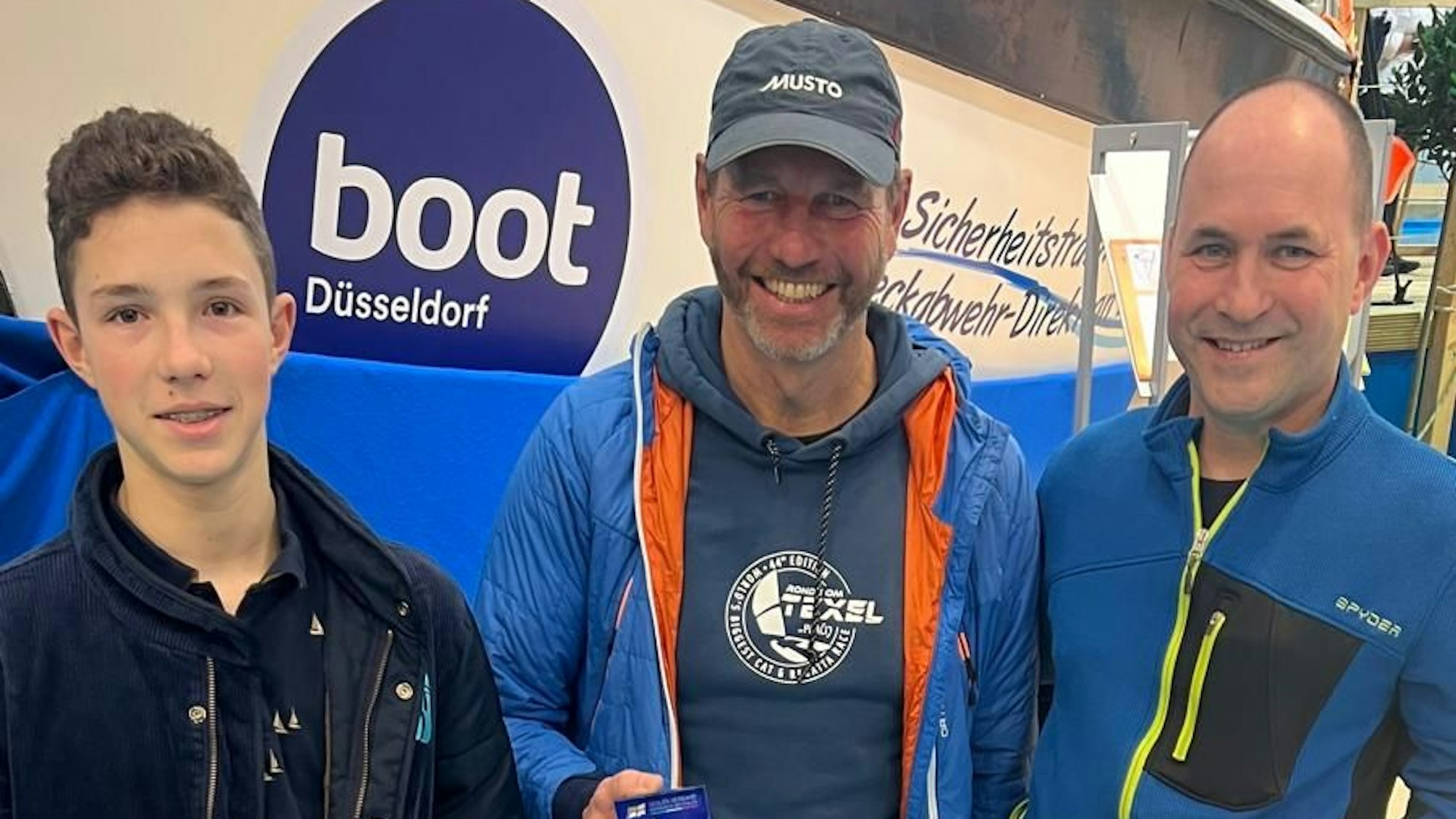Zwei Männer und ein Jugendlicher stehen vor einem Schild der Boot in Düsseldorf und lächeln in die Kamera.