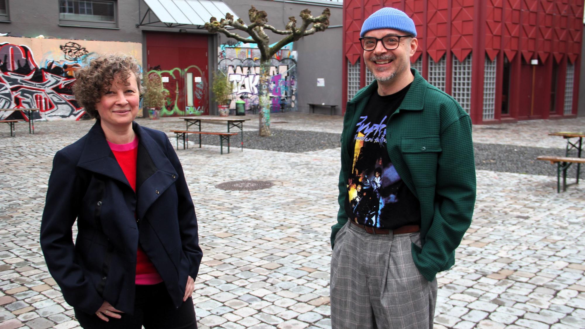 Eine Frau mit Locken und ein Mann mit Brille und blauer Mütze stehen in einem Innenhof.