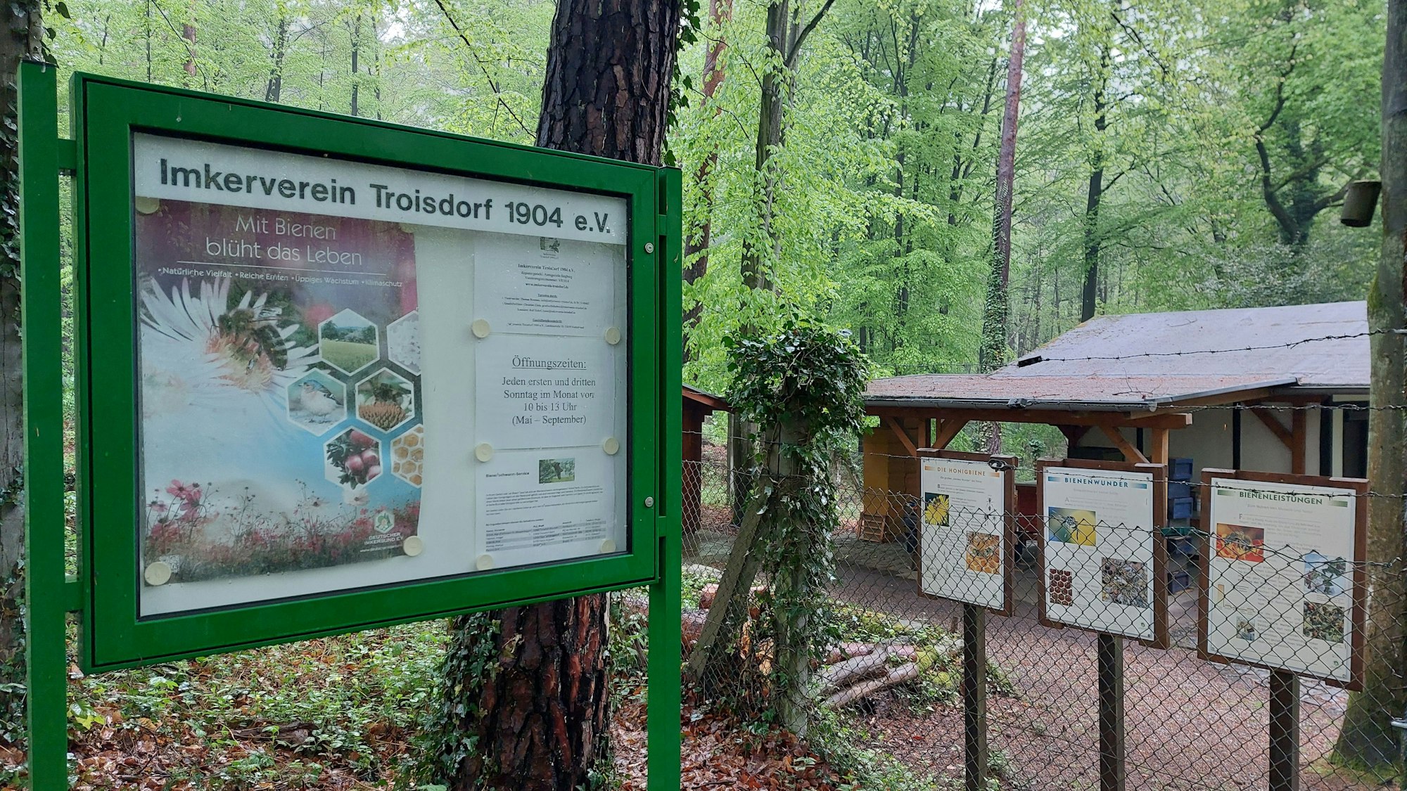 Ein Schaukasten und ein kleines Haus hinter einem Zaun im Wald.