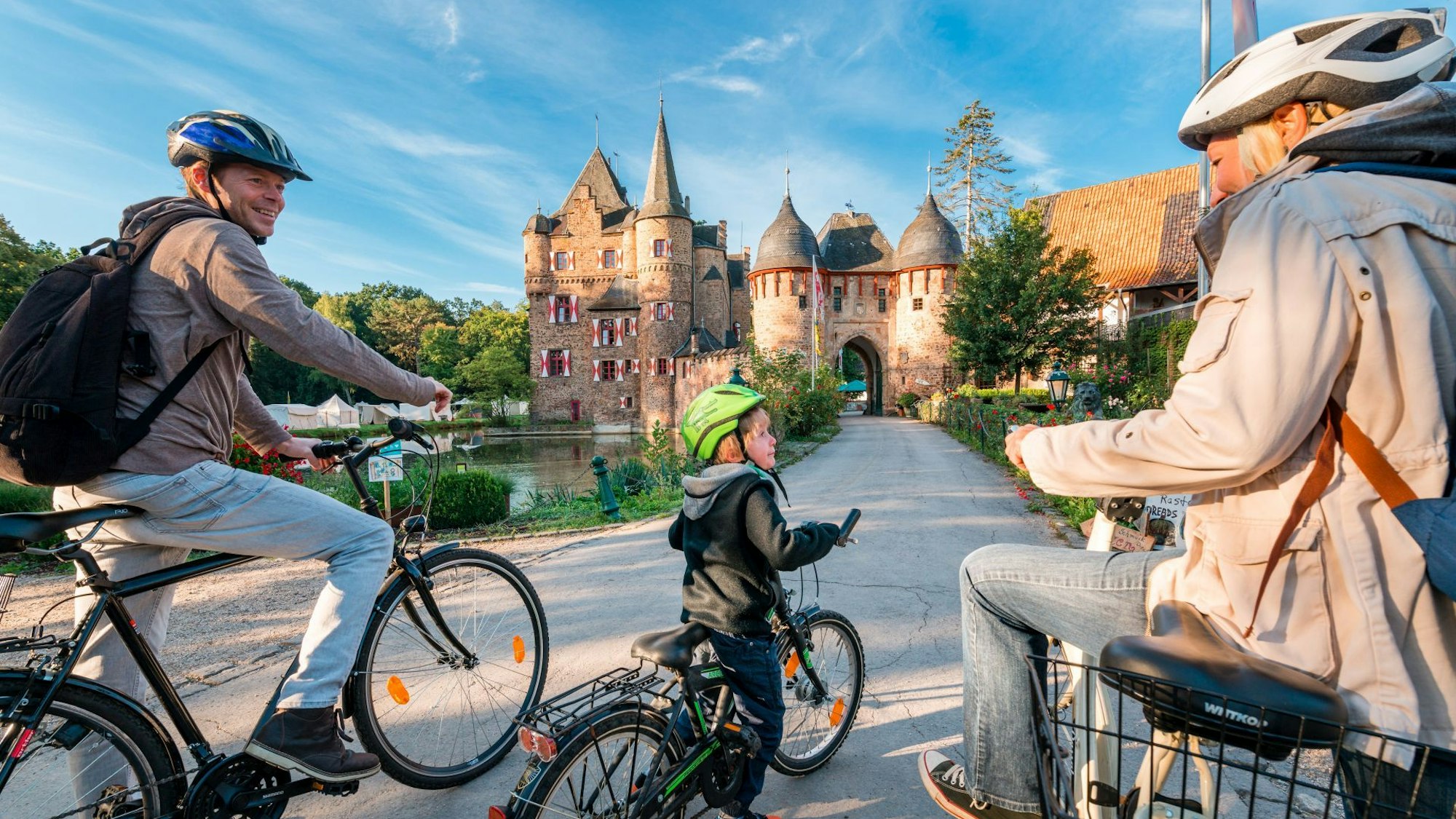 Drei Radfahrer, zwei Erwachsene und ein Kind, vor der Burg Satzvey