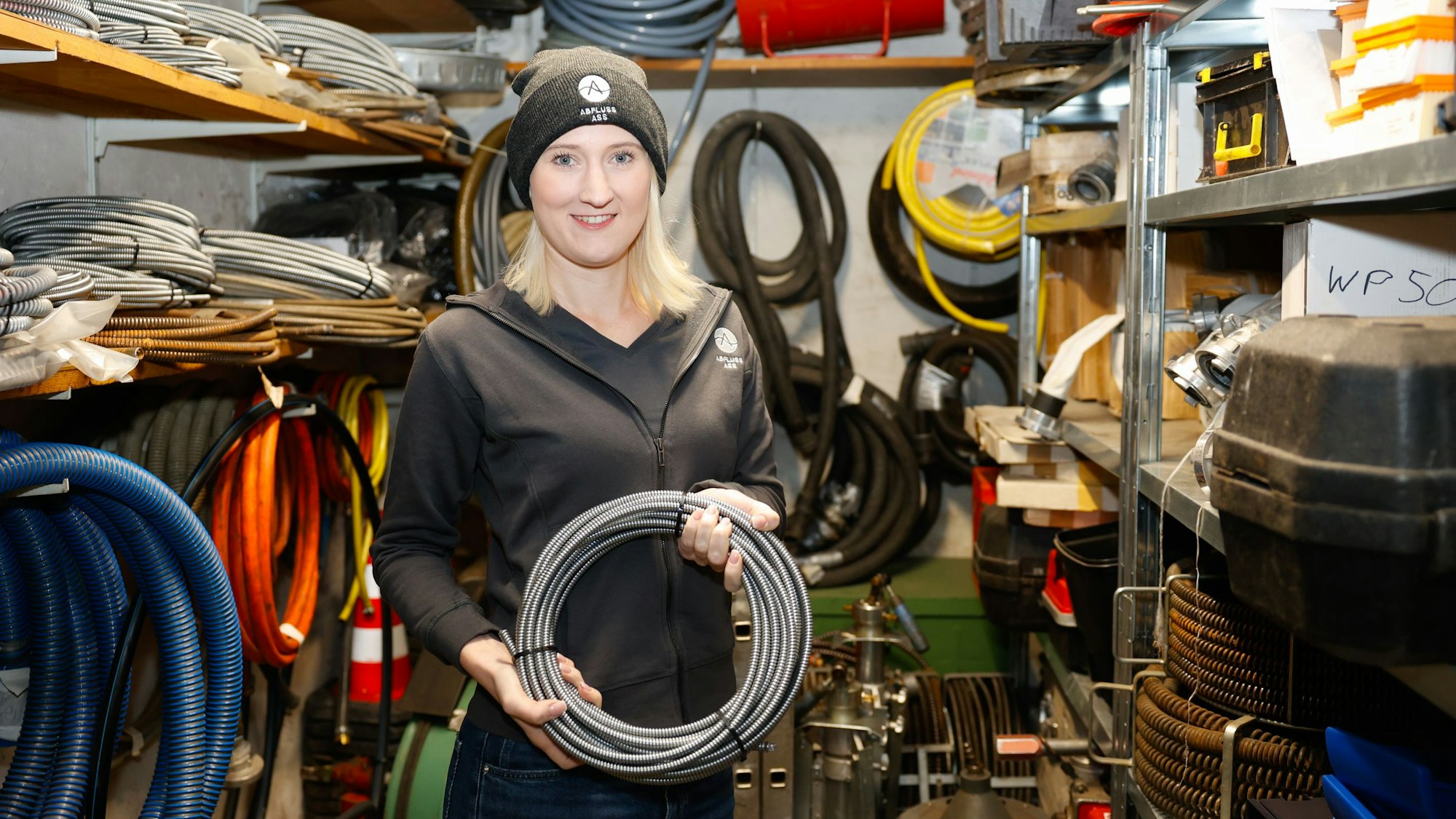 Carina Straub hält ein Kabel in der Hand und steht in einer Werkzeugkammer. Sie führt seit sieben Jahren in dritter Generation die Rohrreinigungsfirma Abfluss ASS Köln.