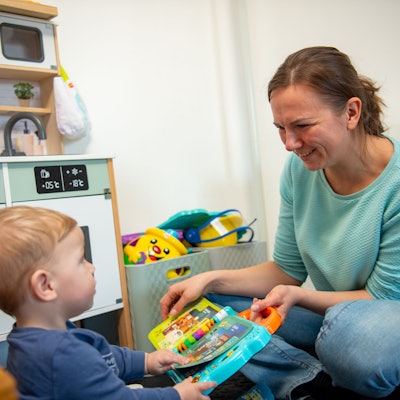 Eine Frau in einem türkisblauen Pullover und Jeans sitzt auf dem Boden und spielt mit einem Kind.