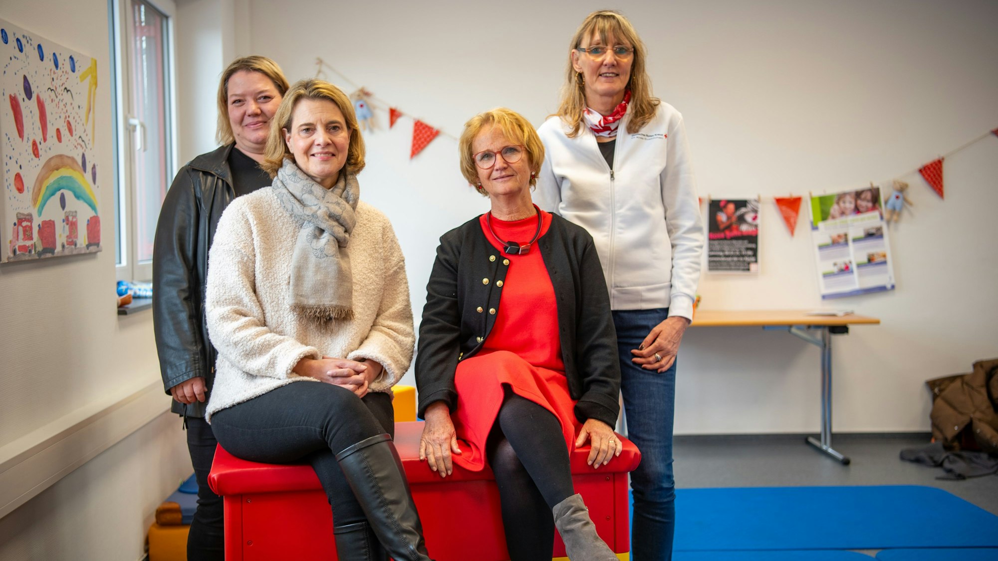 Britta Fünfzig, Leitungsteam AWO-Familienzentrum Frauenberger Straße, (v.l.), Clarissa Lüßem-Meurer, Leitungsteam AWO-Familienzentrum Frauenberger Straße, Miriam Nachtkamp, Leiterin des Müttergesprächskreises und Heike Iven, Leitung Familienbildung DRK, sitzen und stehen auf und an einem bunten Turnkasten. Im Hintergrund sind Turnmatten und bunte Dekoration zu sehen.