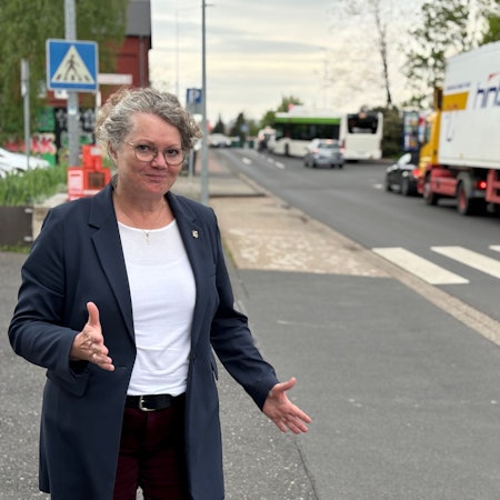 Bürgermeisterin Anna-Katharina Horst steht an einer Straße, an der mehrere Pkw, sowie ein Bus und Lkw stehen. Mit den Händen macht sie eine ausladende Geste.