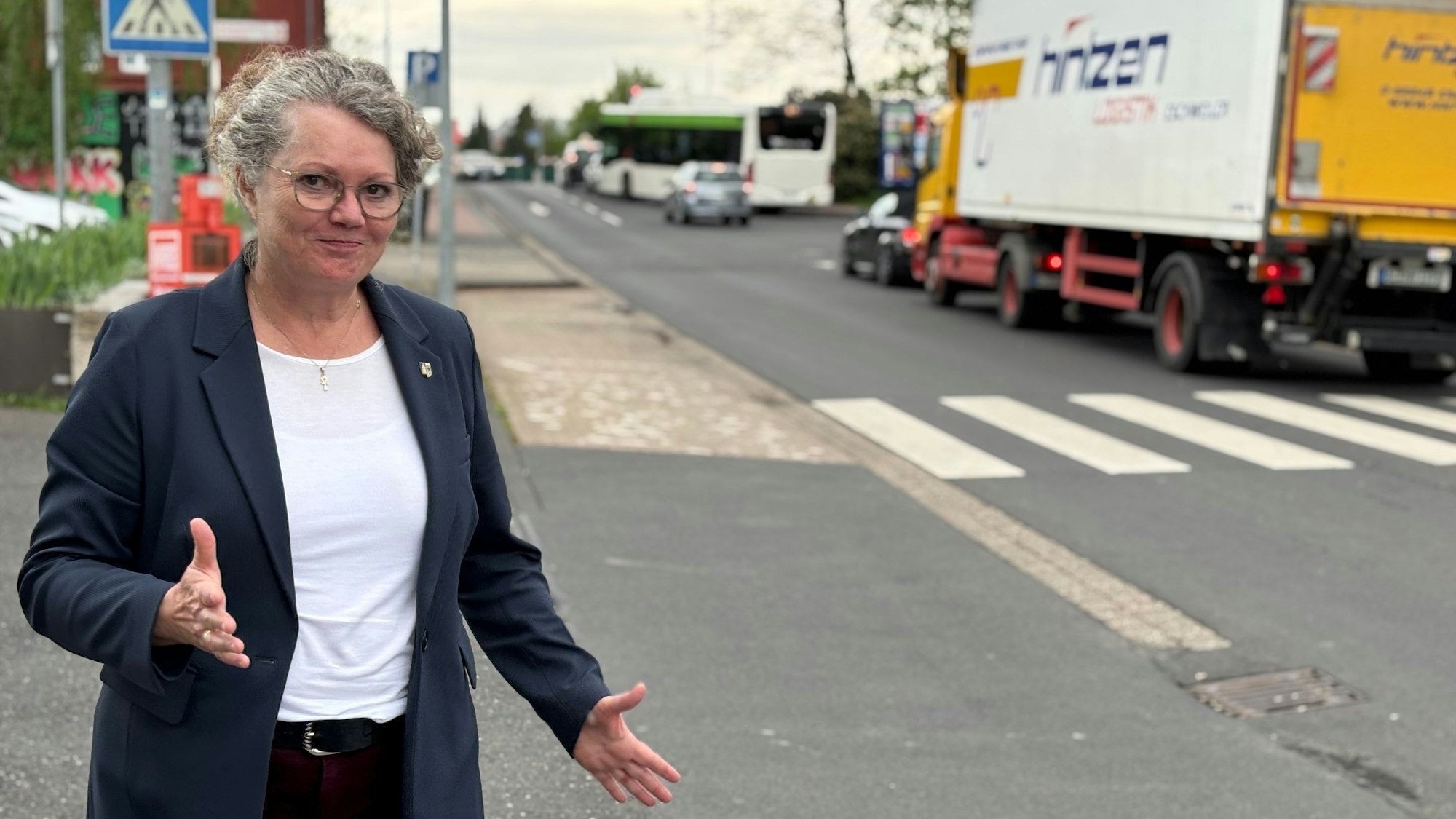 Bürgermeisterin Anna-Katharina Horst steht an einer Straße, an der mehrere Pkw, sowie ein Bus und Lkw stehen. Mit den Händen macht sie eine ausladende Geste.