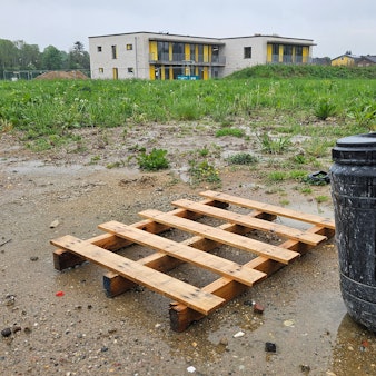 Zu sehen ist eine Holzpalette und zwei schwarze Plastiktonnen vor einer unbebauten Wiese, im Hintergrund steht ein Gebäude.