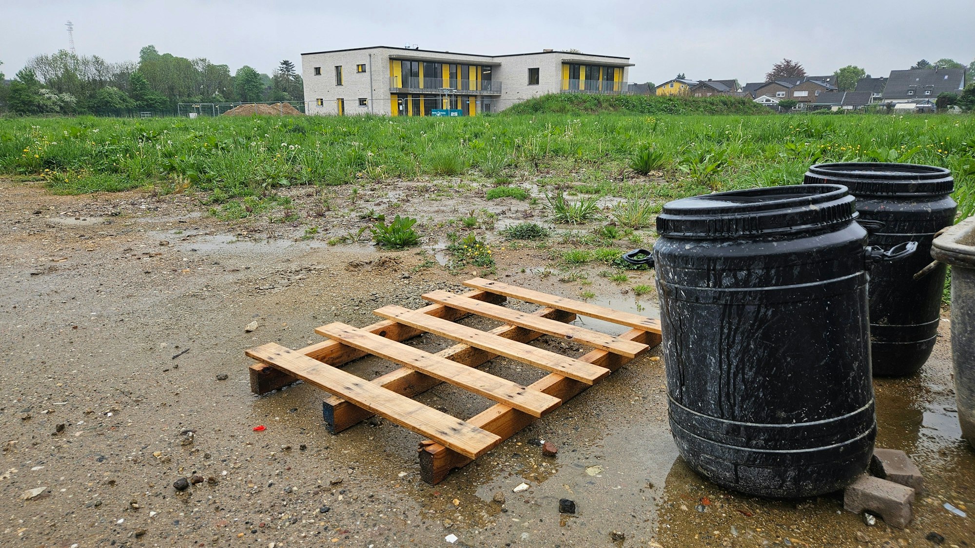 Zu sehen ist eine Holzpalette und zwei schwarze Plastiktonnen vor einer unbebauten Wiese, im Hintergrund steht ein Gebäude.