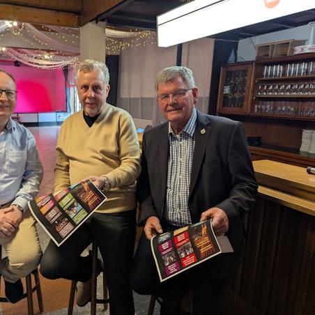 Das Foto zeigt die drei Vorstandsmitglieder auf Barhockern im Saal.