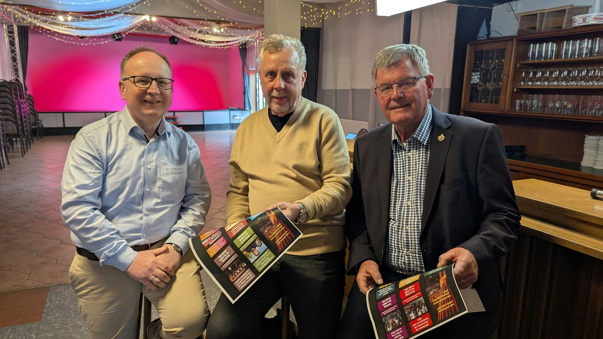 Das Foto zeigt die drei Vorstandsmitglieder auf Barhockern im Saal.