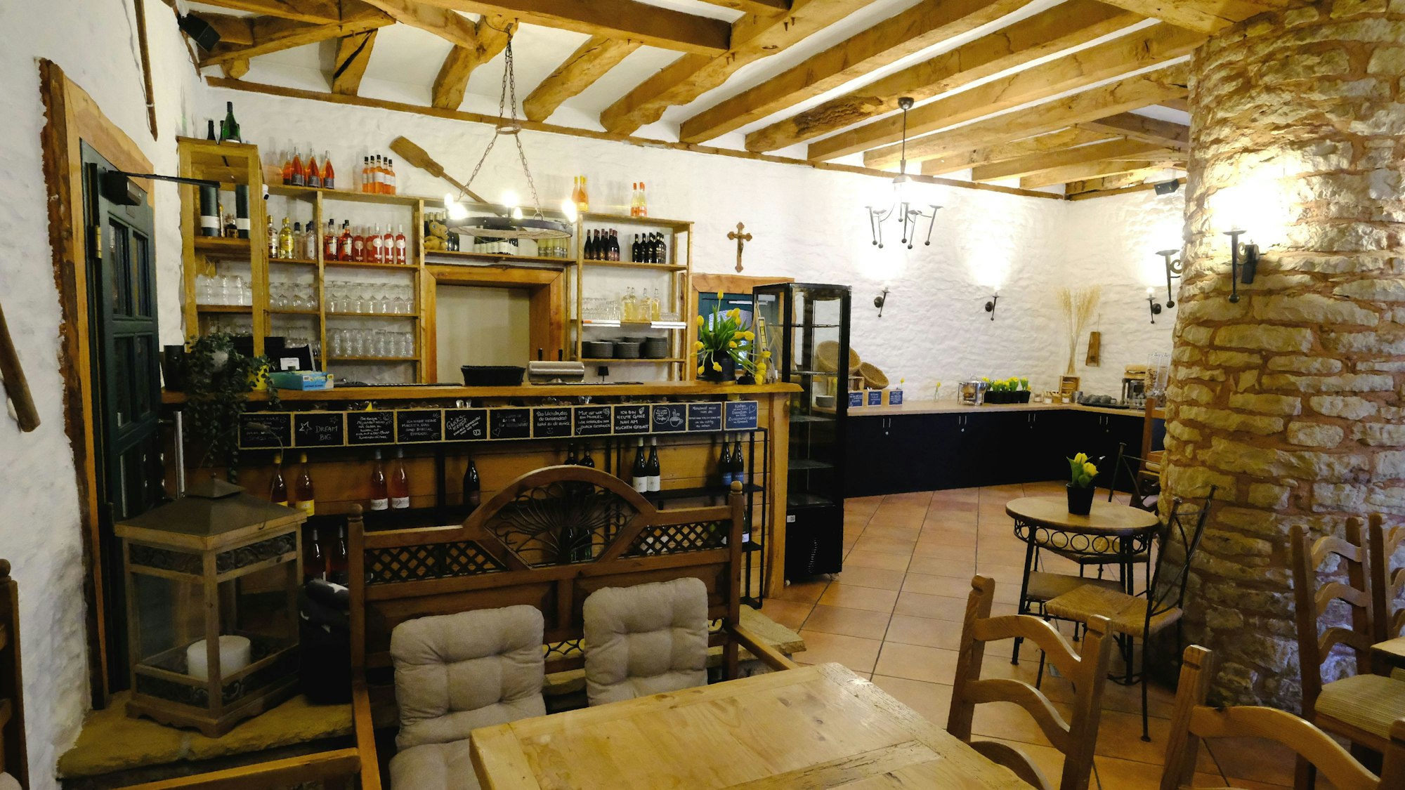 Blick in den Gastraum des Bauerncafés Zur Zehntscheune in Eicks. Der Boden ist gefliest, es gibt eine Decke mit Holzbalken und eine aus Natursteinen gemauerte Säule zwischen den Tischen und Stühlen.