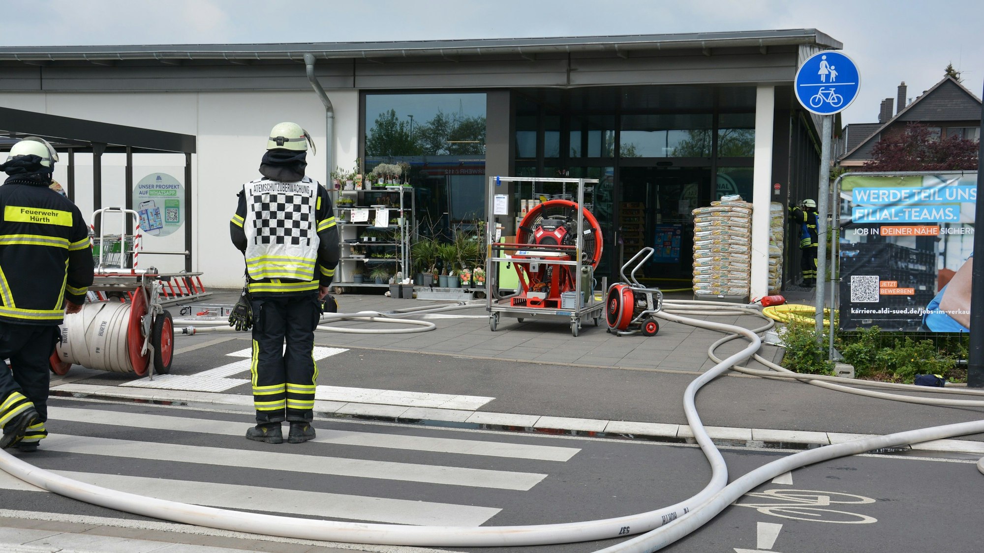 Einsatzkräfte der Feuerwehr mit Schläuchen und einem Großlüfter vor dem Eingang der Aldi-Filiale.