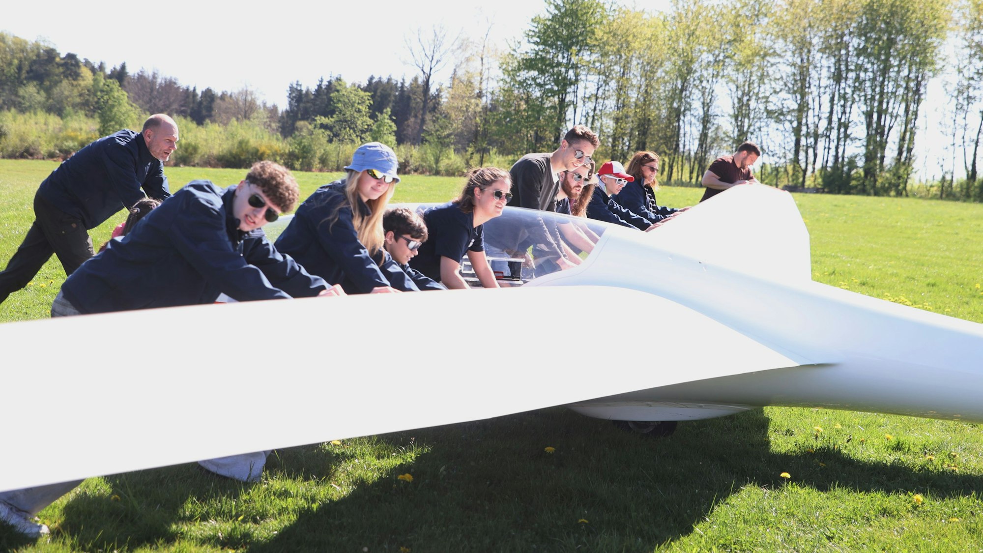 Eine Gruppe von Menschen schiebt ein Segelflugzeug über eine Wiese.