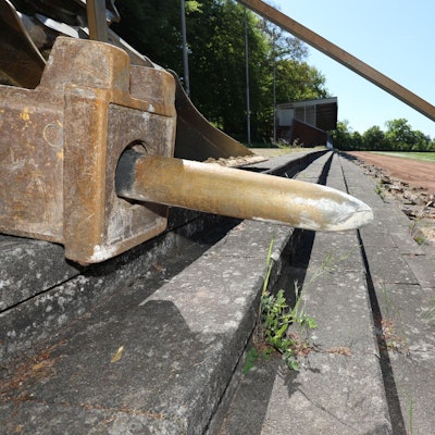 Der überdimensionale Meißel eines Baggers liegt auf Sitzstufen aus Beton, die stark beschädigt sind.