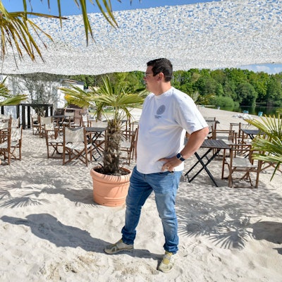 Zu sehen ist Christoph Hausmann, der im weißen Sand vor Töpfen mit Palmen steht.