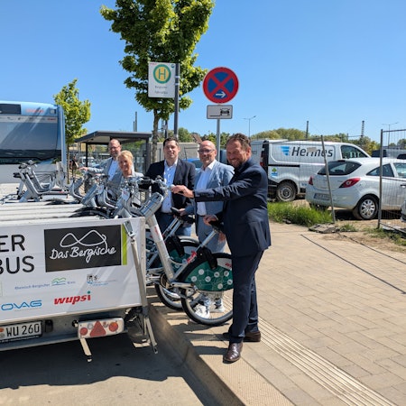 Vier Männer und eine Frau heben Räder von einem Anhänger hinter einem Bus.