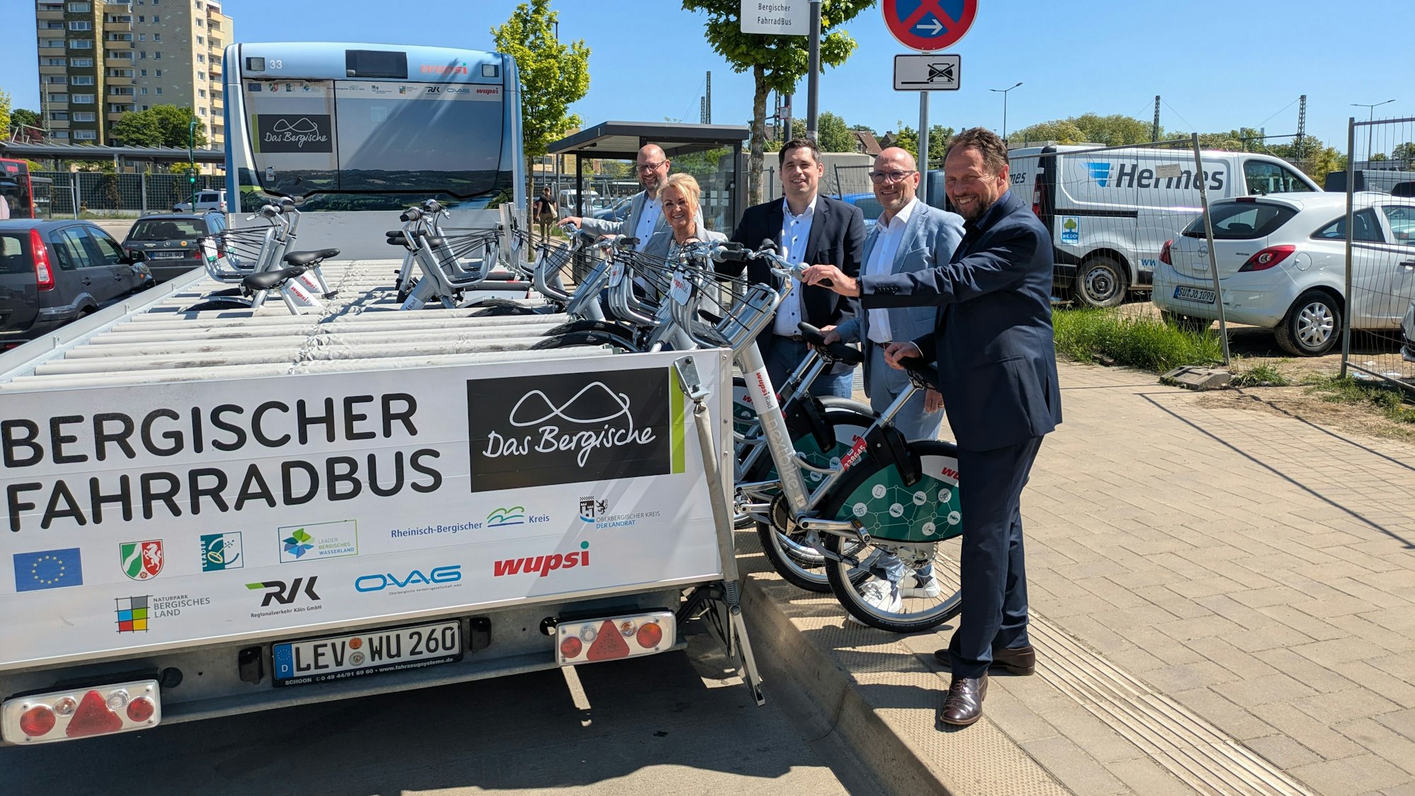 Vier Männer und eine Frau heben Räder von einem Anhänger hinter einem Bus.