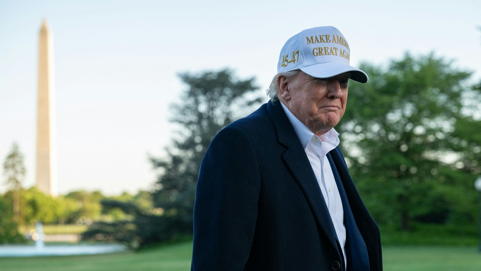 Donald Trump steht vor dem Weißen Haus. Er trägt eine Kappe mit der Aufschrift „Make America Great Again“. Im Hintergrund ist das Washington Memorial zu sehen.