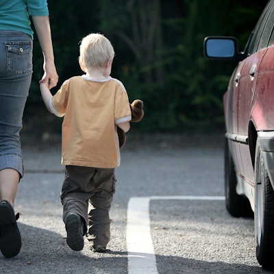 Eine Mutter geht mit ihrem Sohn an der Hand zwischen Autos durch. Das Kind hat ein Kuscheltier im Arm.