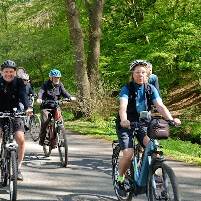 Das Bild zeigt Radfahrerinnen und Radfahrer auf einem asphaltierten Weg, im Hintergrund Bäume und Sträucher.
