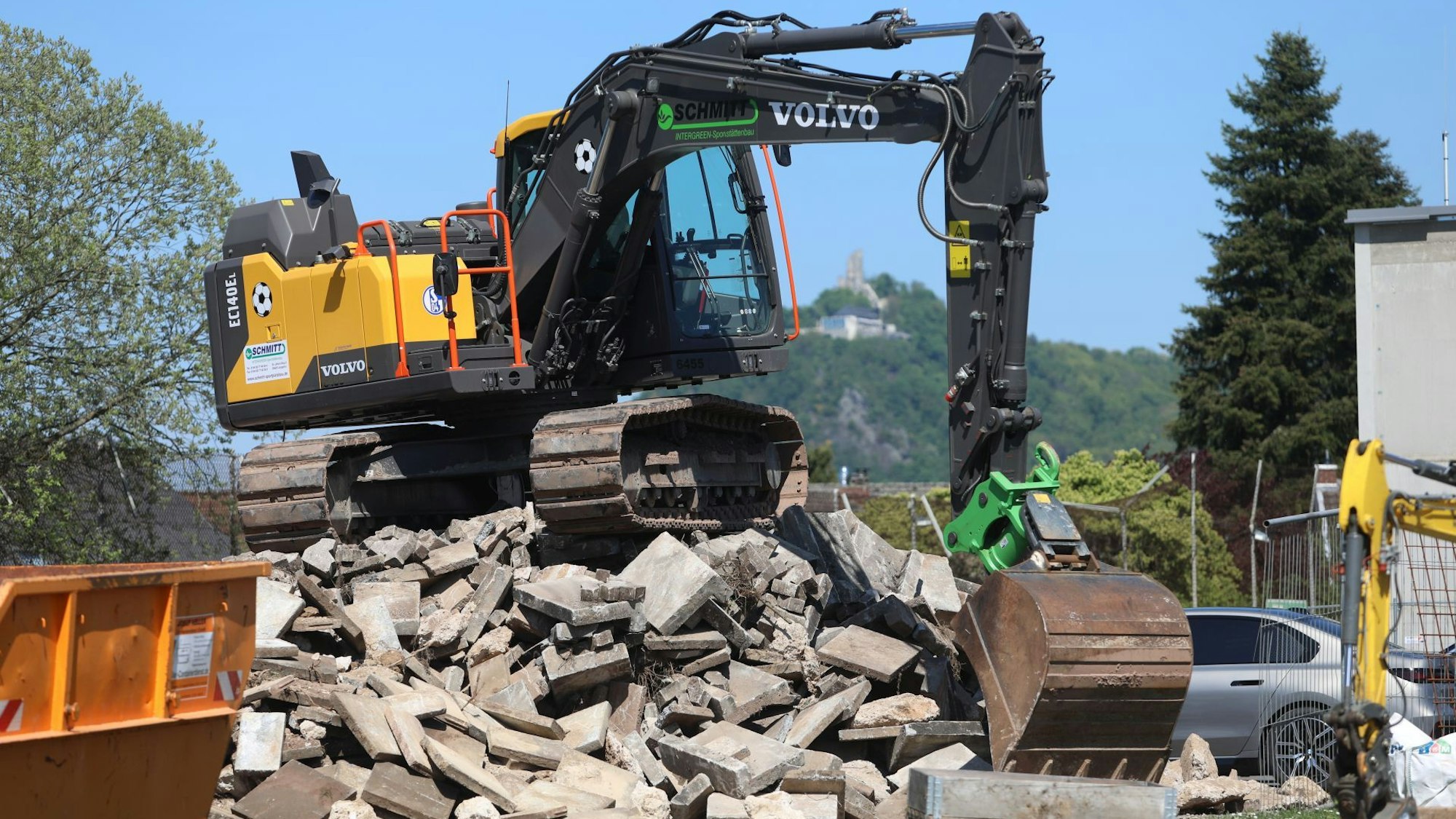 Ein gelber Bagger steht auf einem Schuttberg, im Hintergrund ist der Drachenfels in Königswinter zu erkennen.