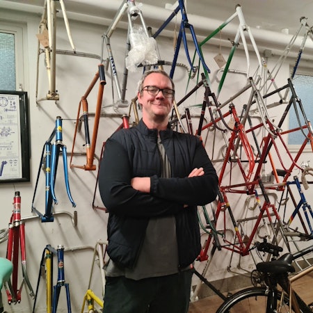 Ein Mann mit Brille und dunklen Klamotten steht vor einer Wand, an der Fahrradrahmen in allen Farben hängen.