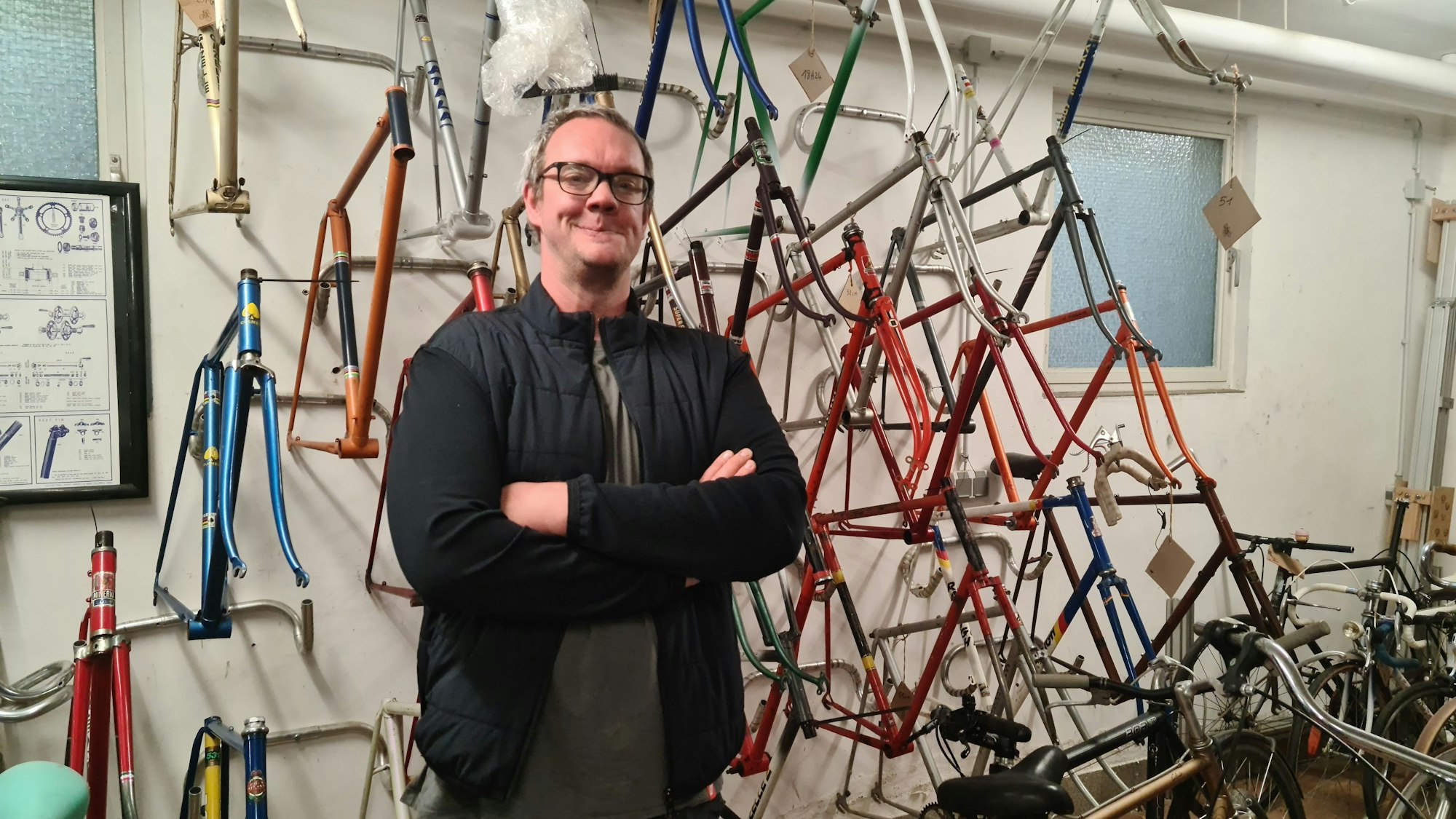 Ein Mann mit Brille und dunklen Klamotten steht vor einer Wand, an der Fahrradrahmen in allen Farben hängen.