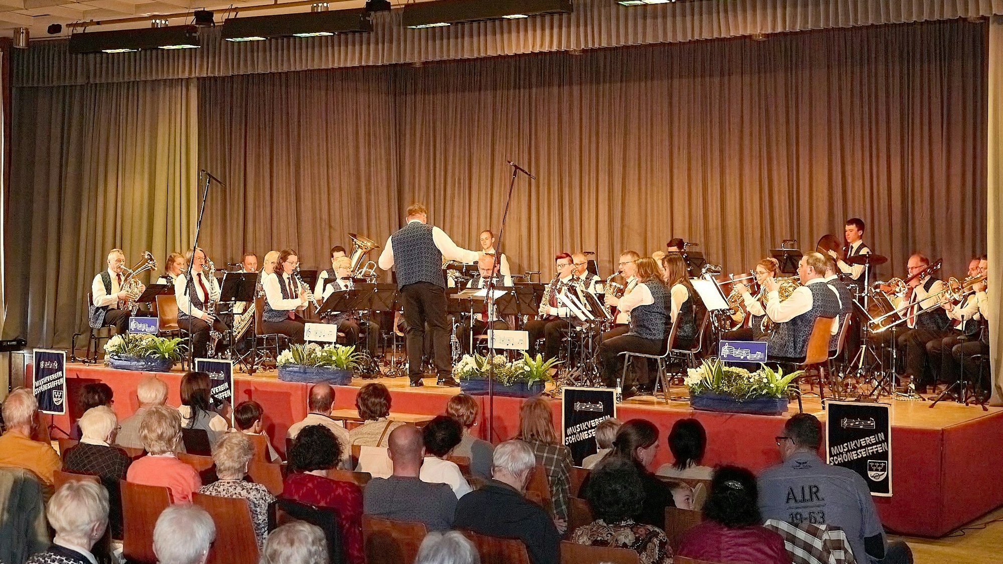 Die Musikerinnen und Musiker spielen im Sitzen auf der Bühne, im Vordergrund das Publikum.