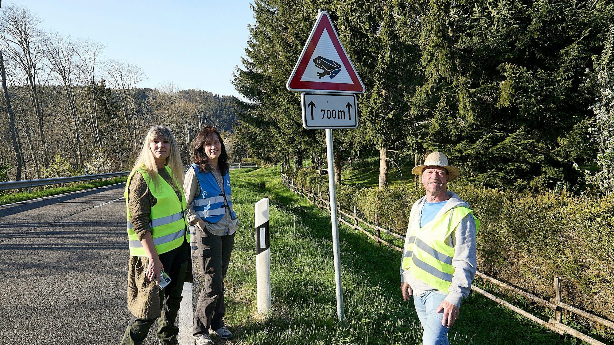 Zwei Frauen und ein Mann tragen Warnwesten. Sie stehen an der Landstraße bei Hellenthal-Wildenburg, an der ein Warnschild mit einer Kröte angebracht ist, das auf die Wanderung der Amphibien hinweist.