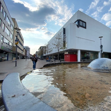 Zu sehen ist das Gebäude der Sinn-Filiale in Brühl und im Vordergrund das Wasser eines Brunnens.
