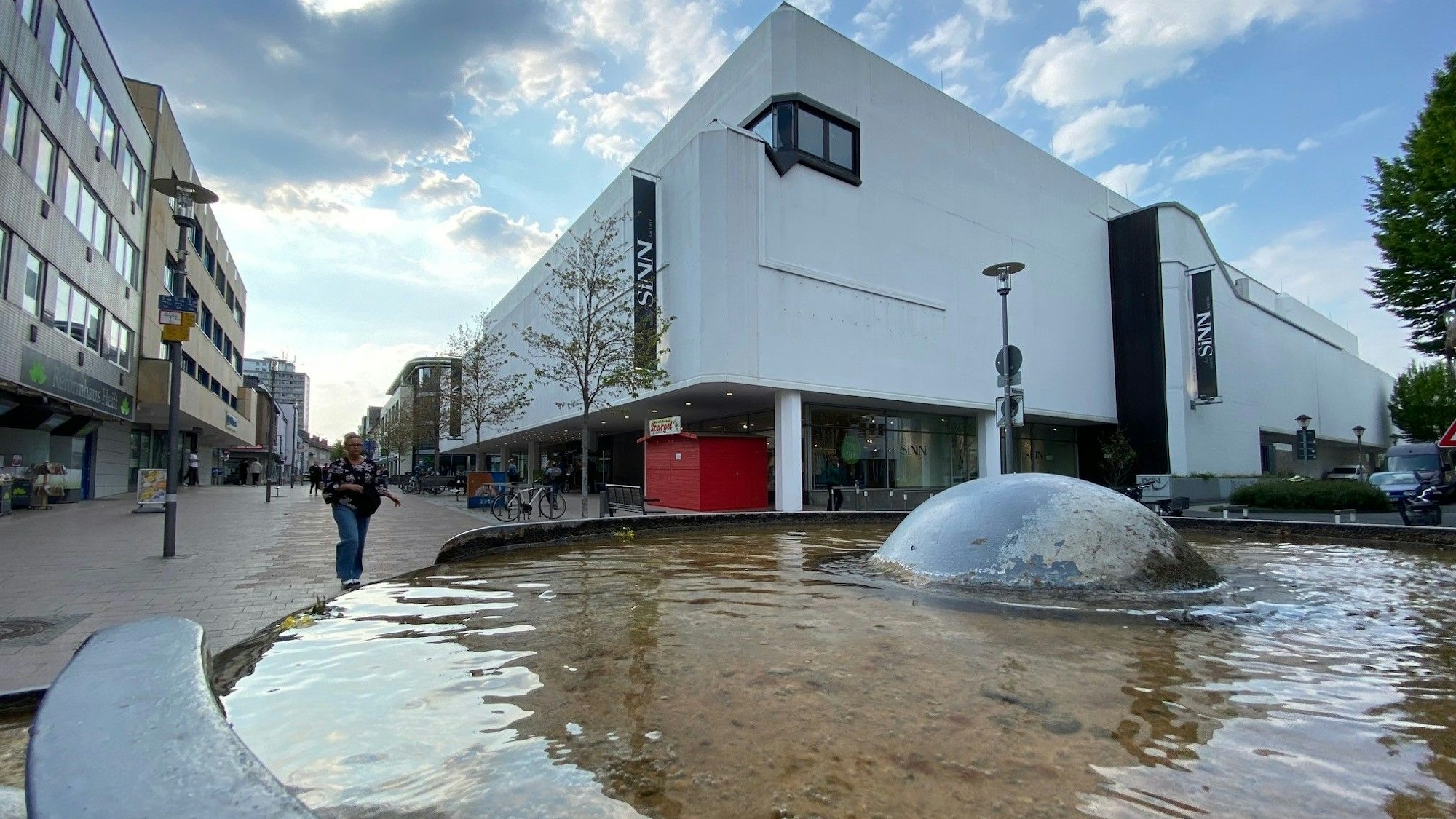 Zu sehen ist das Gebäude der Sinn-Filiale in Brühl und im Vordergrund das Wasser eines Brunnens.