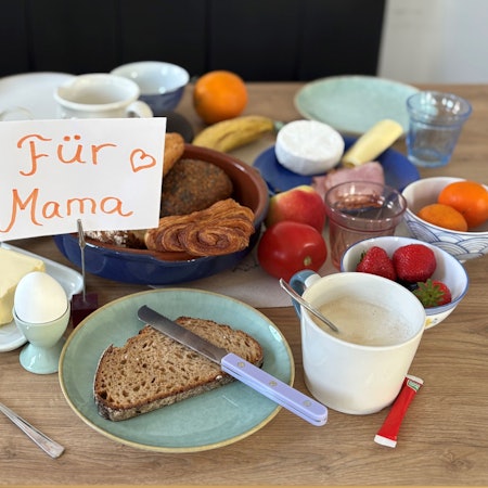 Ein gedeckter Tisch mit Frühstücksspeisen wie Brot, Brötchen, Ei und Obst, dazu ein Schild auf dem "Für Mama" steht.