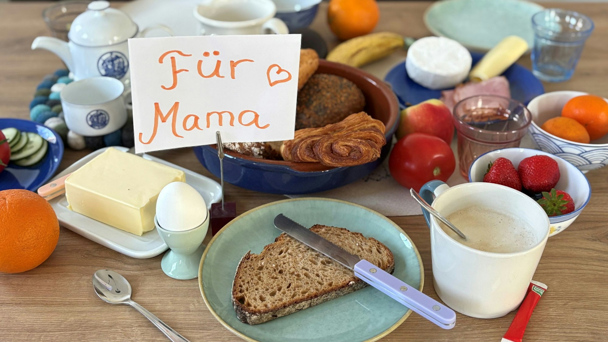 Ein gedeckter Tisch mit Frühstücksspeisen wie Brot, Brötchen, Ei und Obst, dazu ein Schild auf dem "Für Mama" steht.